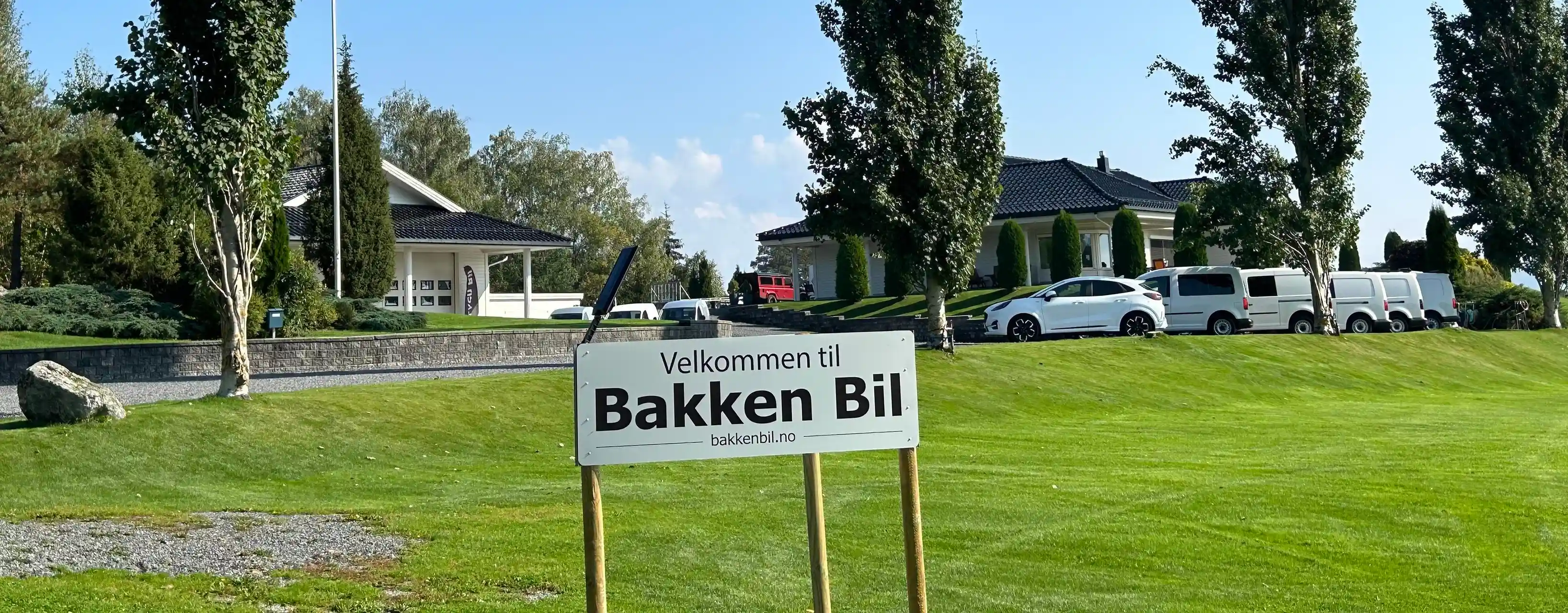 Et skilt der det ønskes velkommen til Bakken Bil i forgrunnen, med en vakker eiendom i bakgrunn der man kan se mange biler stilt opp på rekke og rad
